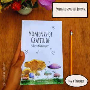 Gratitude journal titled "Moments of Gratitude" includes hand illustrated mushrooms on the front cover with little doodle hearts rising up. Paperback journal with a black and white interior.