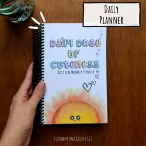 Daily planner titled Daily Dose of Cuteness daily and monthly planner has pastel colored lettering in the title with a smiling sunshine rising up from the bottom edge of the spiralbound and laminated cover.