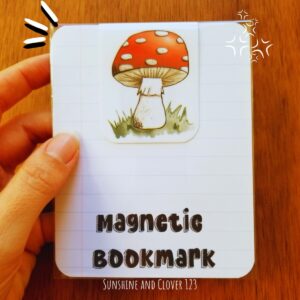 Magnetic bookmark with hand illustrated red mushroom and white spots on green grass.